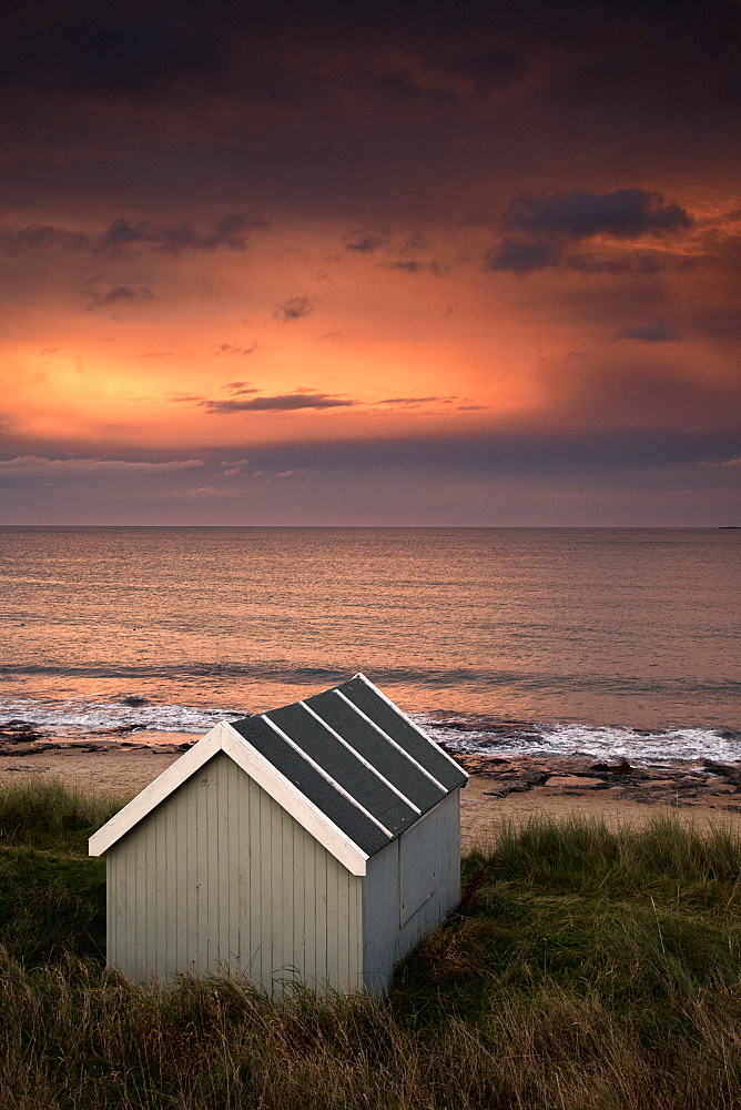 A Small Building Along The Water With A Pink Sky At Sunset, Bamburgh, Northumberland, England