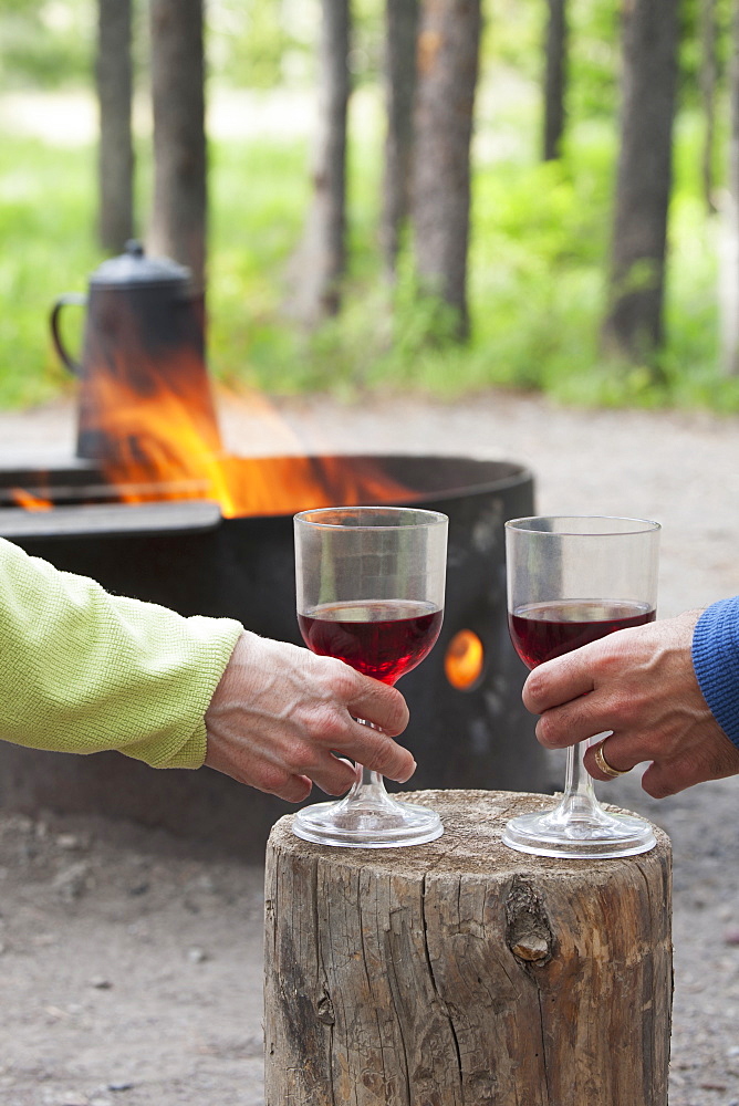 A Couple Holding Wine Glasses On A Block Of Wood With A Fire Pit Burning In A Campground, Waterton, Alberta, Canada