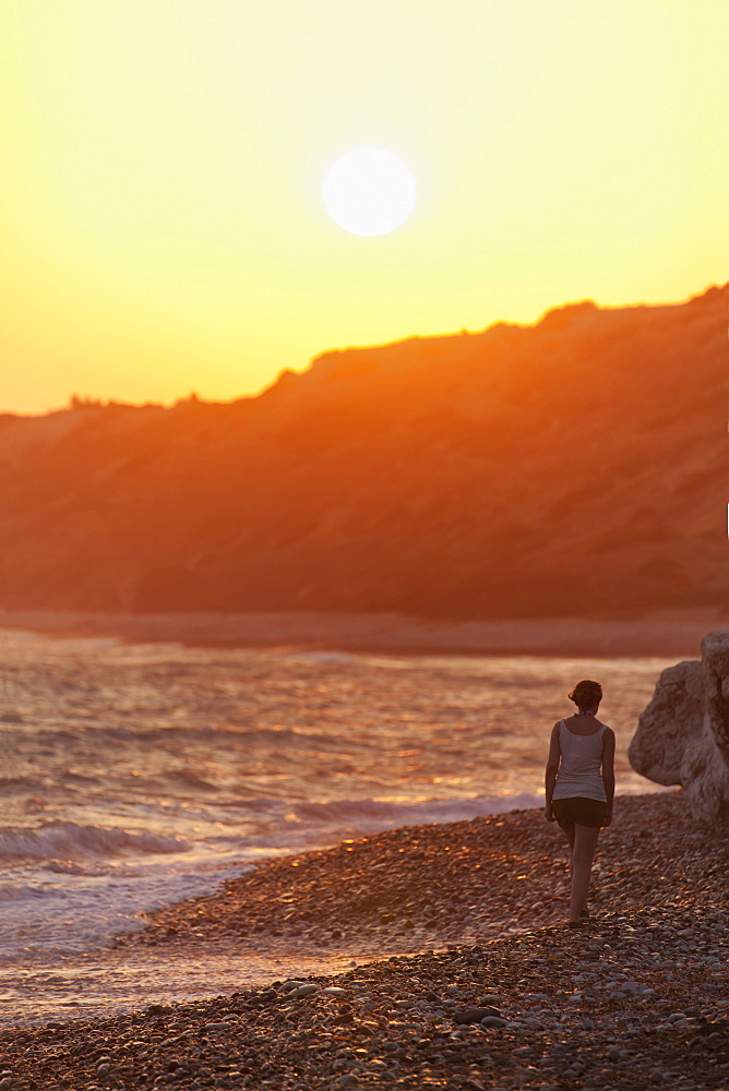 A woman walks along the rock beach at the water's edge at sunset, Aphrodite beach, cyprus