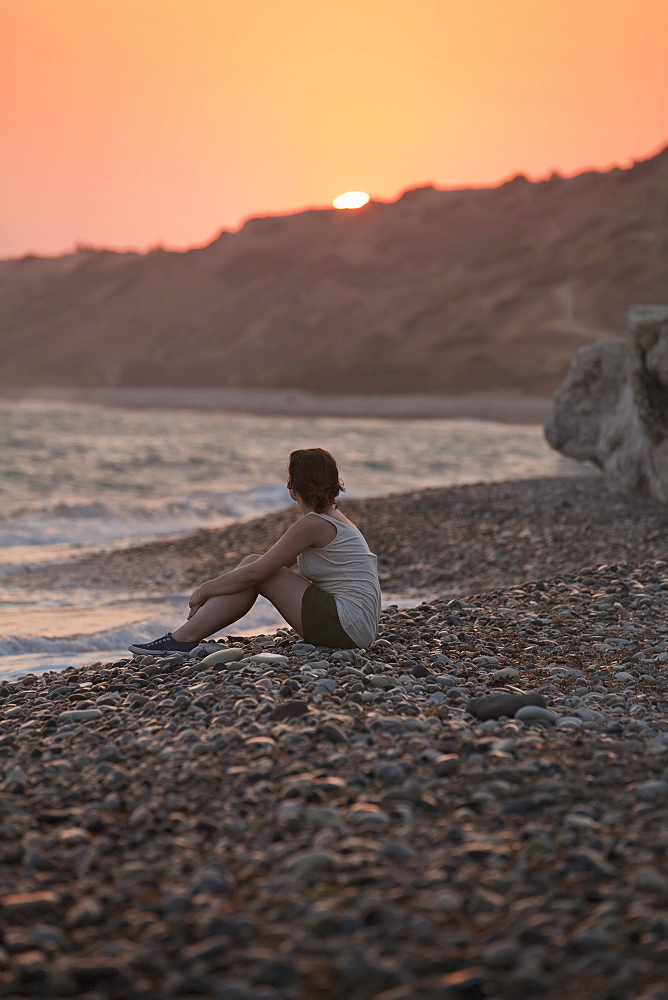 A woman sits on the rock beach looking out to the ocean at sunset, Aphrodite bay, cyprus