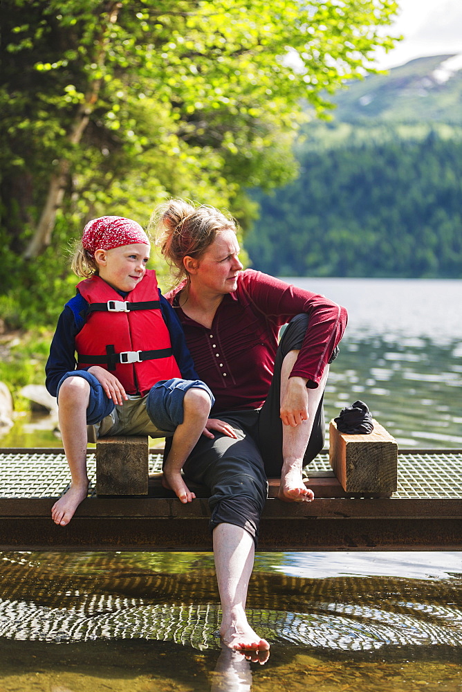 A Mother And Daughter Sits On A Dock Looking Out Over Byers Lake, Soaking Their Bare Feet In The Water, Denali State Park, Alaska, United States Of America