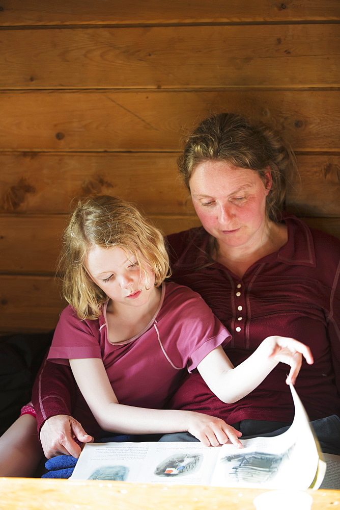 A Mother And Daughter Sitting Together Reading A Book, Leaning Against A Wooden Cabin Wall, Alaska, United States Of America