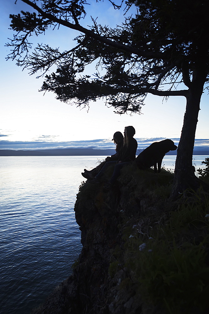 A Couple With A Dog Sit On A Ridge At The Water's Edge Looking Out Over Halibut Cove, Kachemak Bay, Kenai Peninsula, Alaska, United States Of America