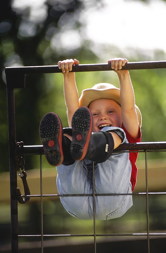 Agriculture - A young farm girl wearing a cowboy hat and rubber boots swings on a metal gate, hanging by her hands and legs and laughing / Northwest Missouri, USA.