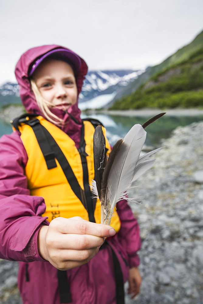 Young Girl In Yellow Lifejacket Displaying Some Bird Feathers Standing In Front Of Shoup Glacier And Chugach Mountains, Shoup Bay State Marine Park, Prince William Sound, Valdez, Alaska, United States Of America