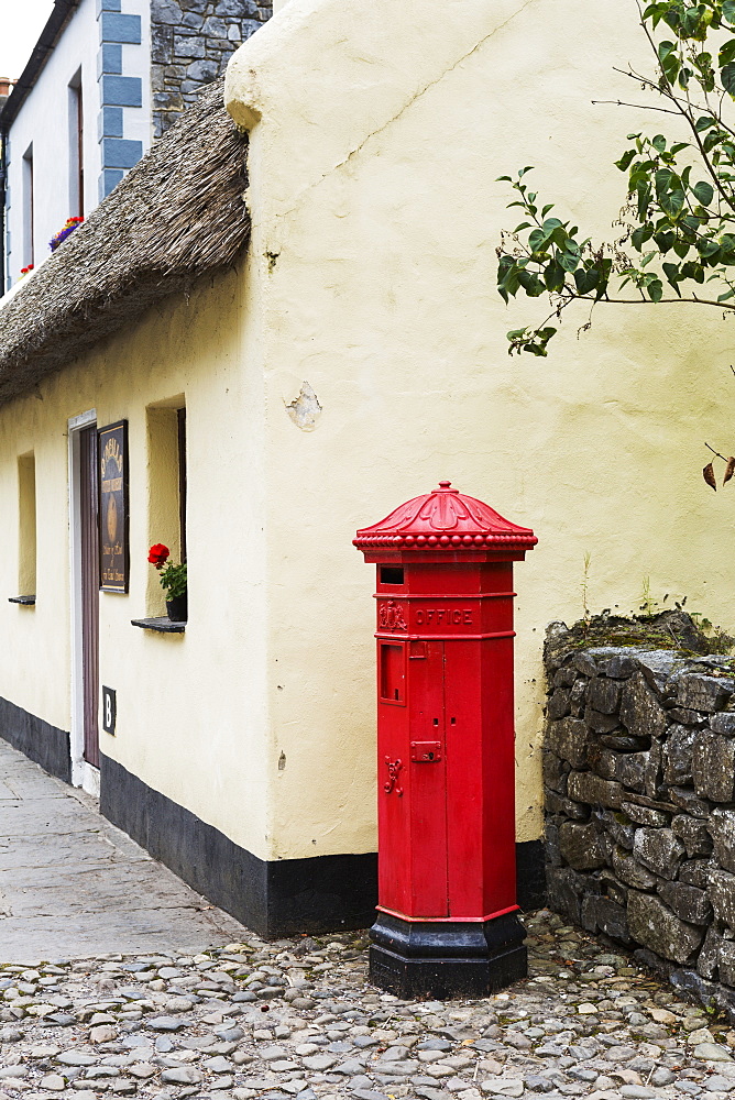 Red Mailbox Standing On Cobblestone Next To Stone Fence With Thatched Roof Building In Background, Bunratty, County Clare, Ireland