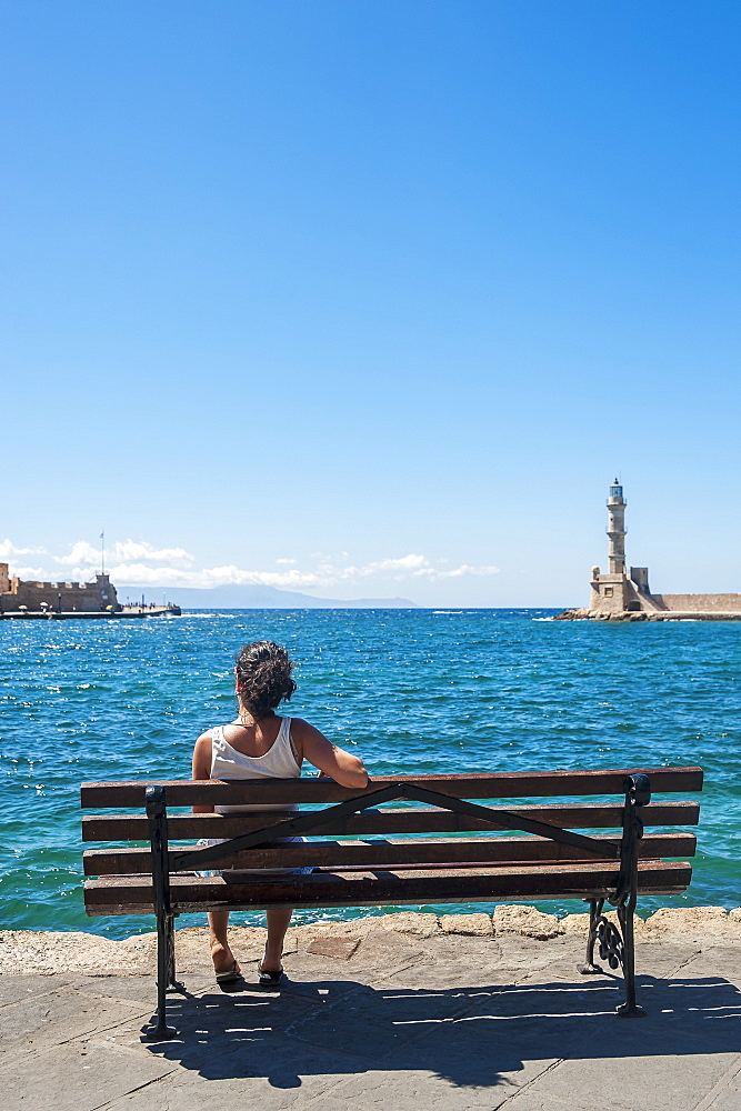 A Woman Sits On A Bench Overlooking The Harbour And Lighthouse, Chania, Crete, Greece