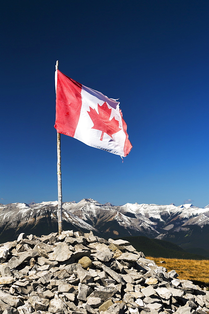 Well Worn Canadian Flag On Wooden Branch Propped In A Pile Of Rocks On Top Of A Mountain With Mountain Range And Blue Sky In The Background, Alberta, Canada