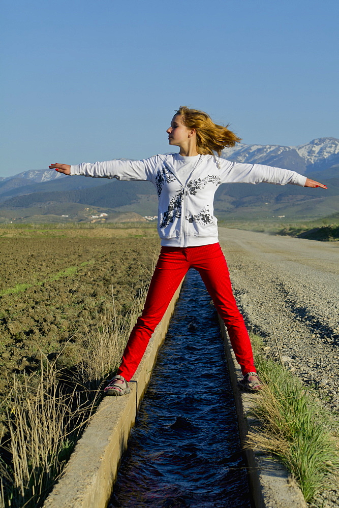 Teenage Girl Standing Over A Small Canal Of Water With Arms Outstretched, Andalusia, Spain