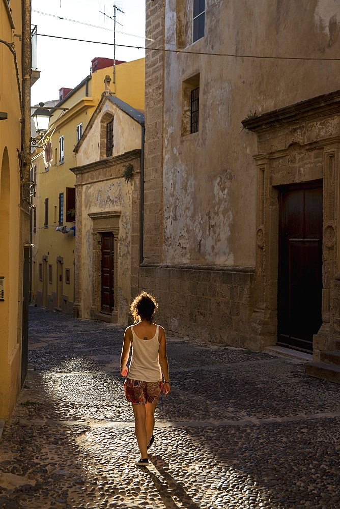 Girl Walking Down A Cobblestone Alley Between Buildings, Alghero, Sardinia, Italy