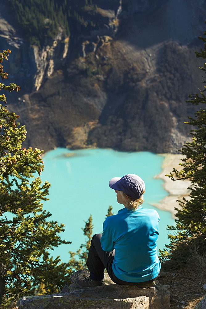 Female Hiker Sitting On Rock High Above Intense Blue Coloured Mountain Lake With Mountain Cliffs In The Background, Banff National Park, Alberta, Canada