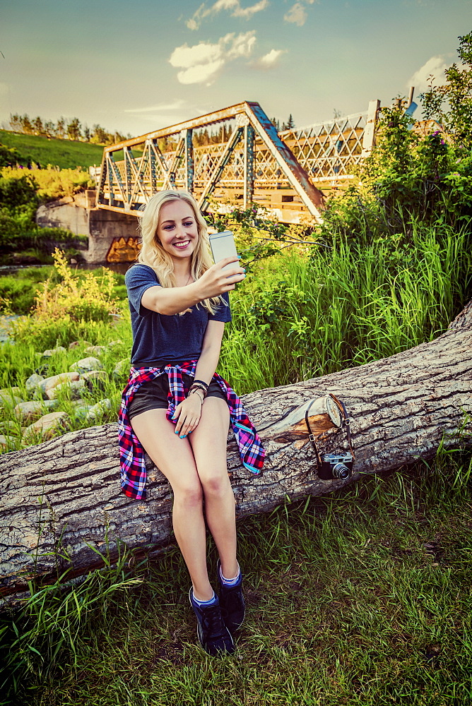 A young woman sits on a log in a park taking a self-portrait with her cell phone with a bridge and river in the background, Edmonton, Alberta, Canada