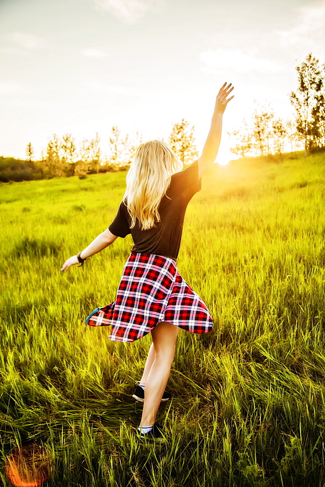 A young woman with long blond hair twirls and runs freely in a grass field in a park at sunset, Edmonton, Alberta, Canada