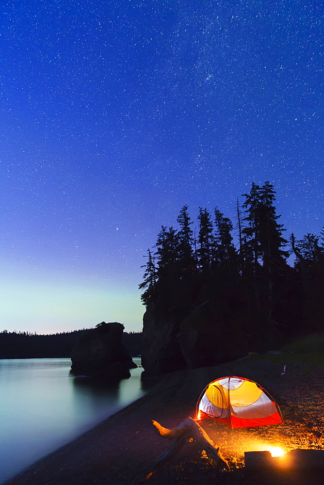 A Glowing Tent On A Beach Overlooks Green Aurora Borealis Reflected In The Tranquil Ocean Water, Hesketh Island, Homer, Alaska, United States Of America