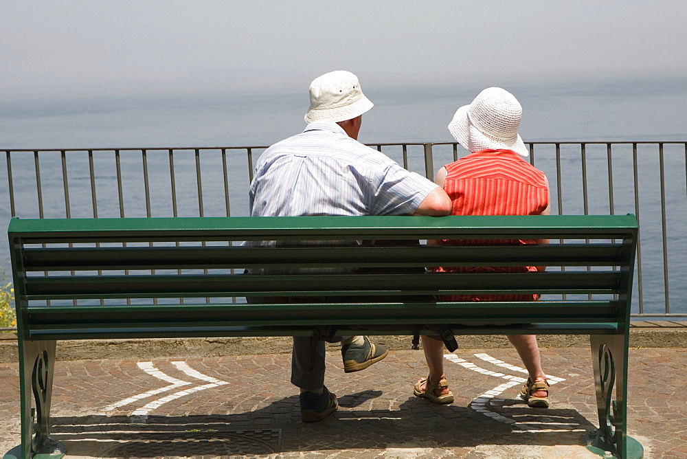 Rear view of a couple sitting on a bench at seaside, Via Aniello Califano, Bay of Naples, Sorrento, Sorrentine Peninsula, Naples Province, Campania, Italy