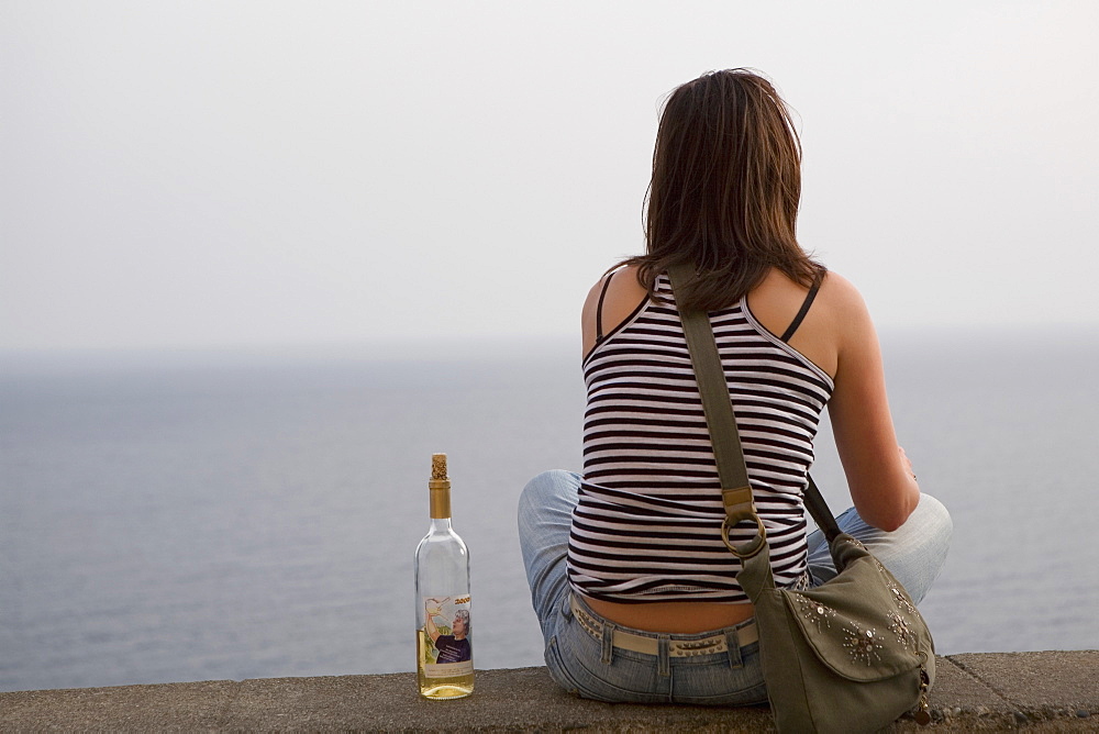 Rear view of a woman sitting at seaside with a wine bottle, Corniglia, Cinque Terre, La Spezia, Liguria, Italy