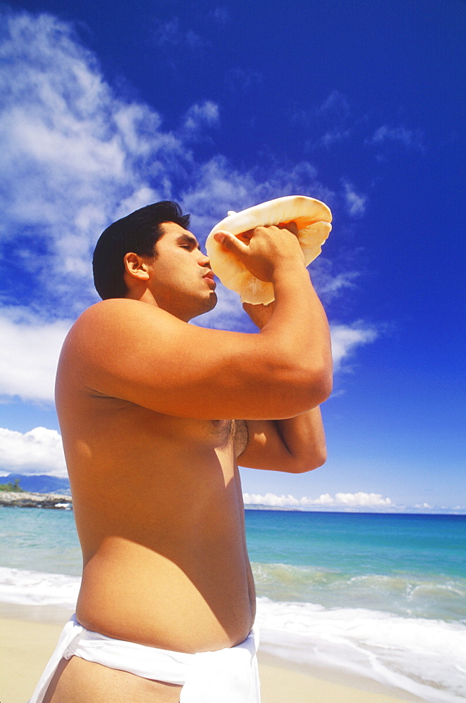 Side profile of a young man blowing a conch shell on the beach, Hawaii, USA