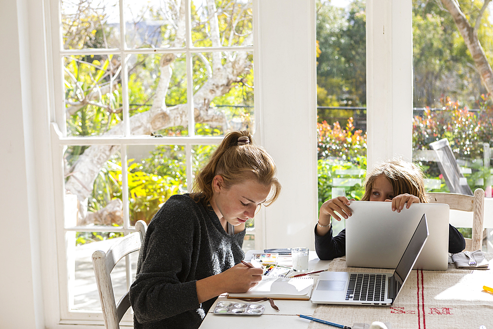 Teenage girl painting with watercolours at a table, and a boy on a laptop