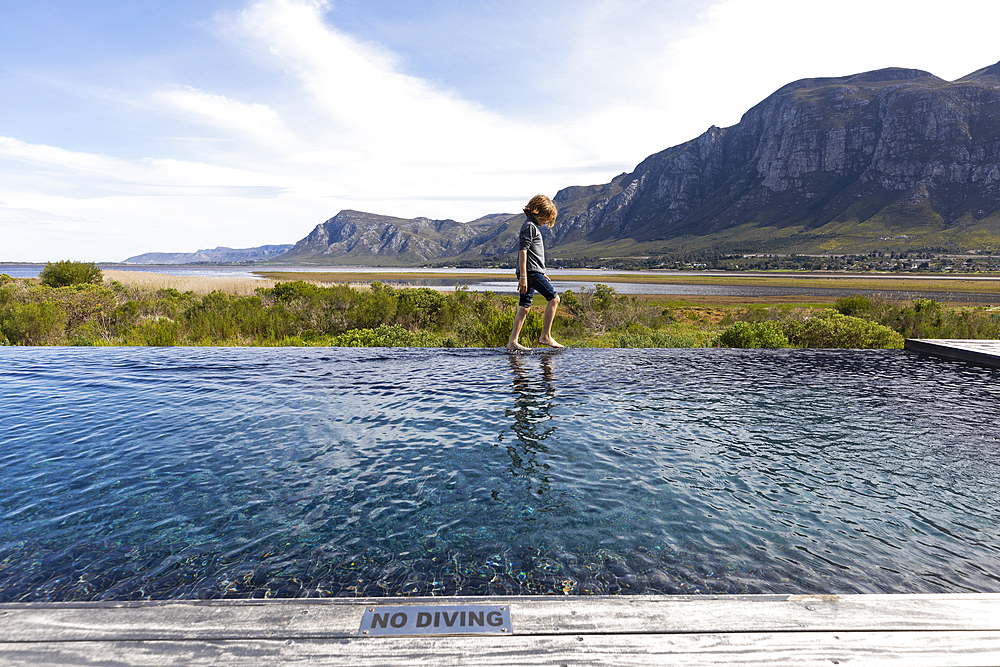 Eight year old boy walking around the edge of an infinity pool, a mountain backdrop