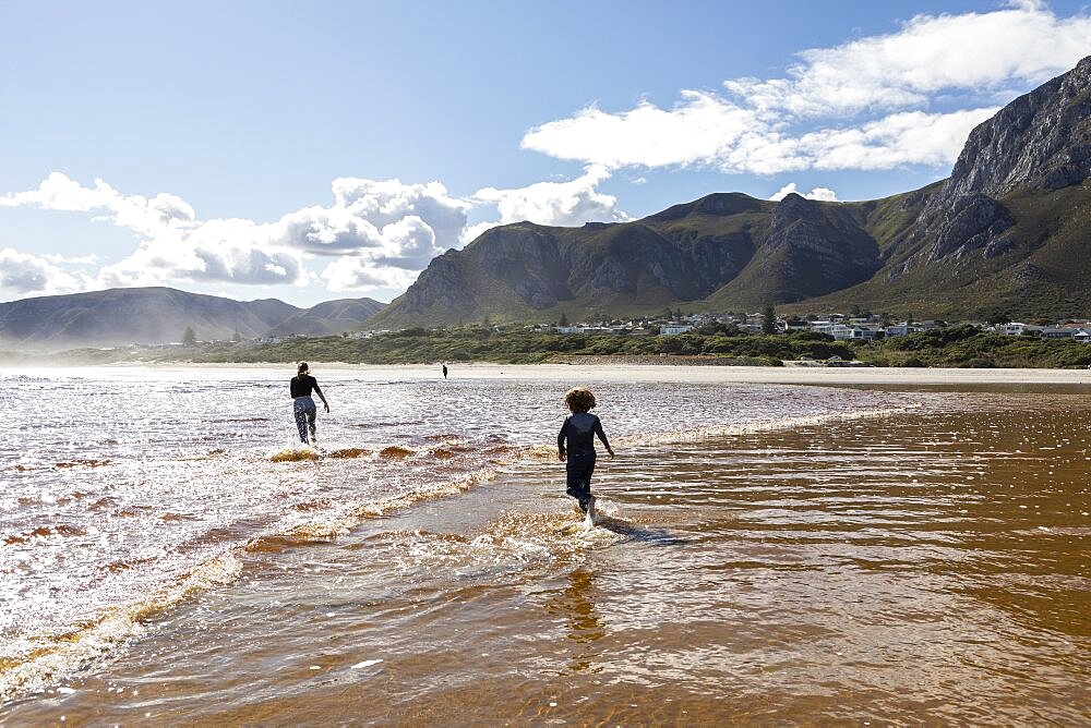 Teenage girl and a boy running through shallow water on a wide sandy beach, Grotto Beach, Western Cape, South Africa