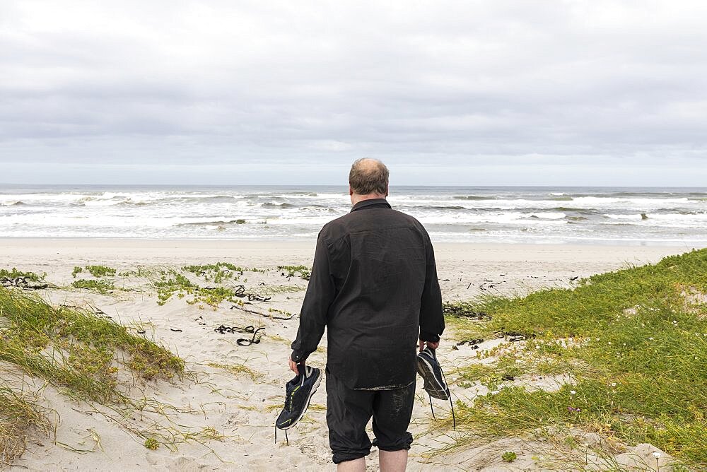 A mature man walking across a beach carrying his shoes in his hand, Grotto Beach, Western Cape, South Africa