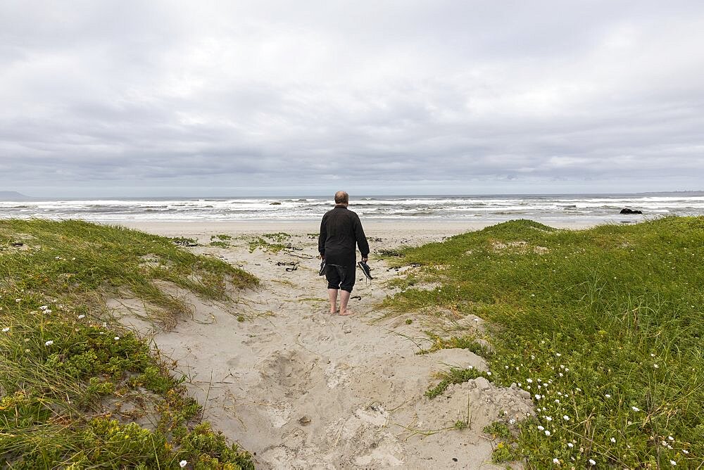 A mature man walking across a beach carrying his shoes in his hand, Grotto Beach, Western Cape, South Africa