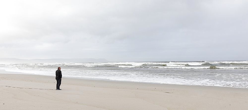 A man walking across sand to the water's edge on a beach, overcast day and surf waves breaking on shore, Grotto Beach, Western Cape, South Africa