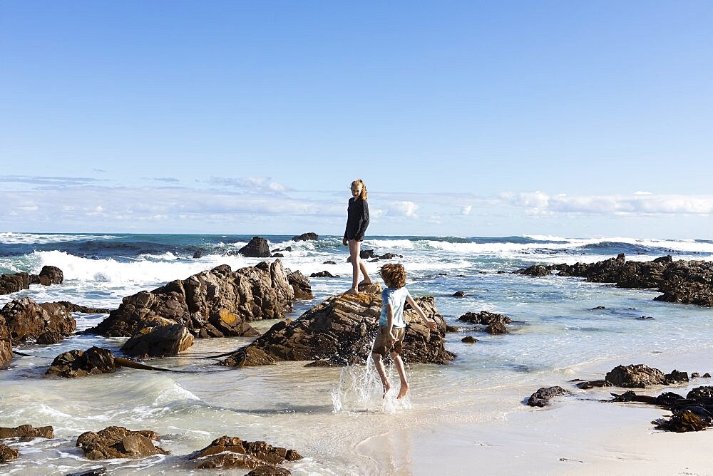 Two children exploring on a sandy beach with jagged rocks and waves breaking, , Pearly Beach, Gansbaai, Western Cape, South Africa