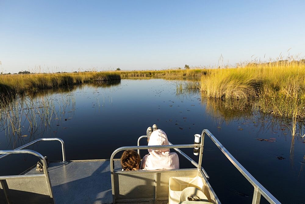 Two children seated on a boat, looking out over calm waters and flat landscape, Okavango Delta, Botswana