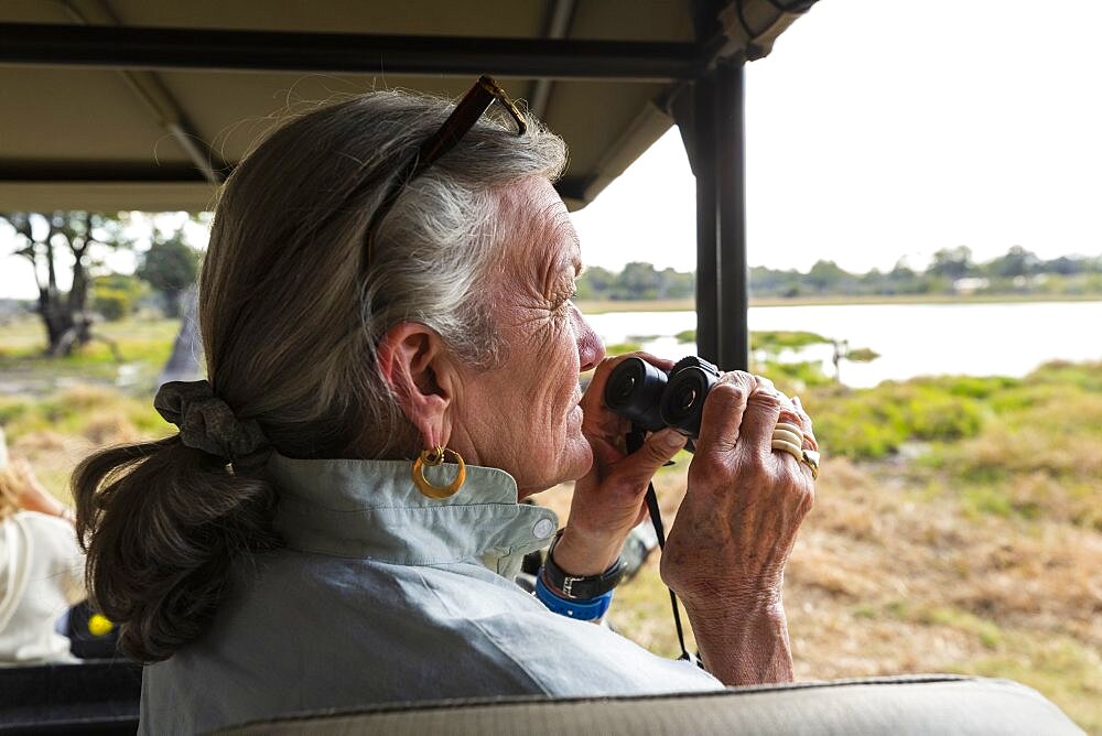 Senior woman using binoculars, sitting in a safari vehicle, looking out over marshes and waterway, Okavango Delta, Botswana