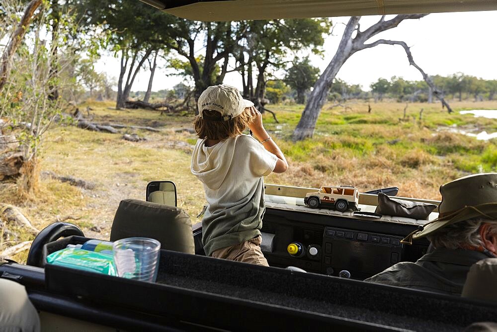 Young boy using binoculars standing up in a safari jeep, looking over the landscape, Okavango Delta, Botswana