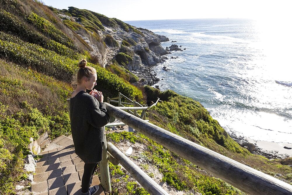 Teenage girl standing on a walkway on the cliffs above a beach, Walker Bay Nature Reserve, South Africa