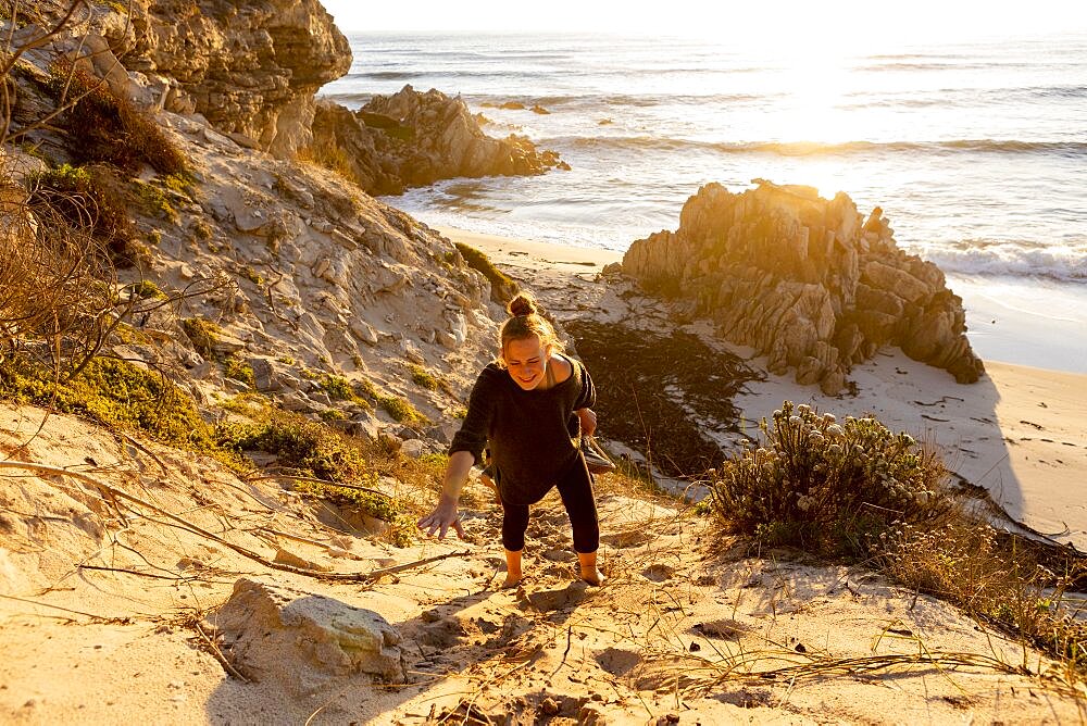 Teenage girl climbing up a very steep sandy slope above a beach, Walker Bay Nature Reserve, South Africa