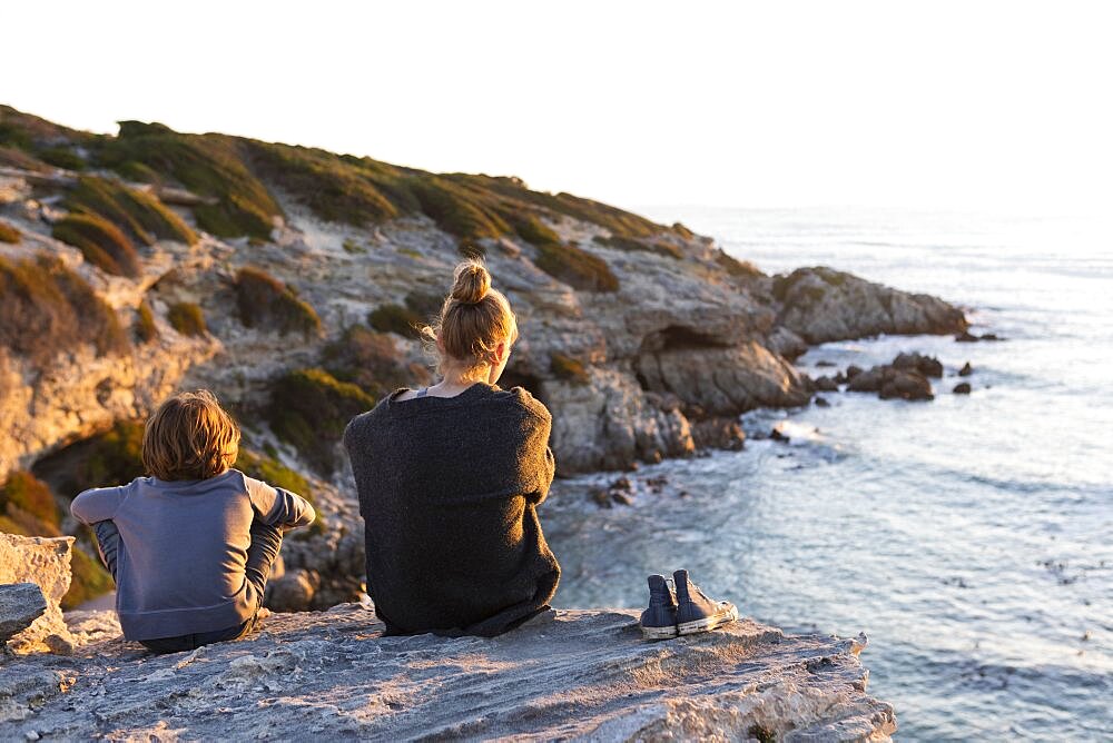 Teenage girl and young boy sitting on rocks looking over the sea at sunset, Walker Bay Nature Reserve, South Africa