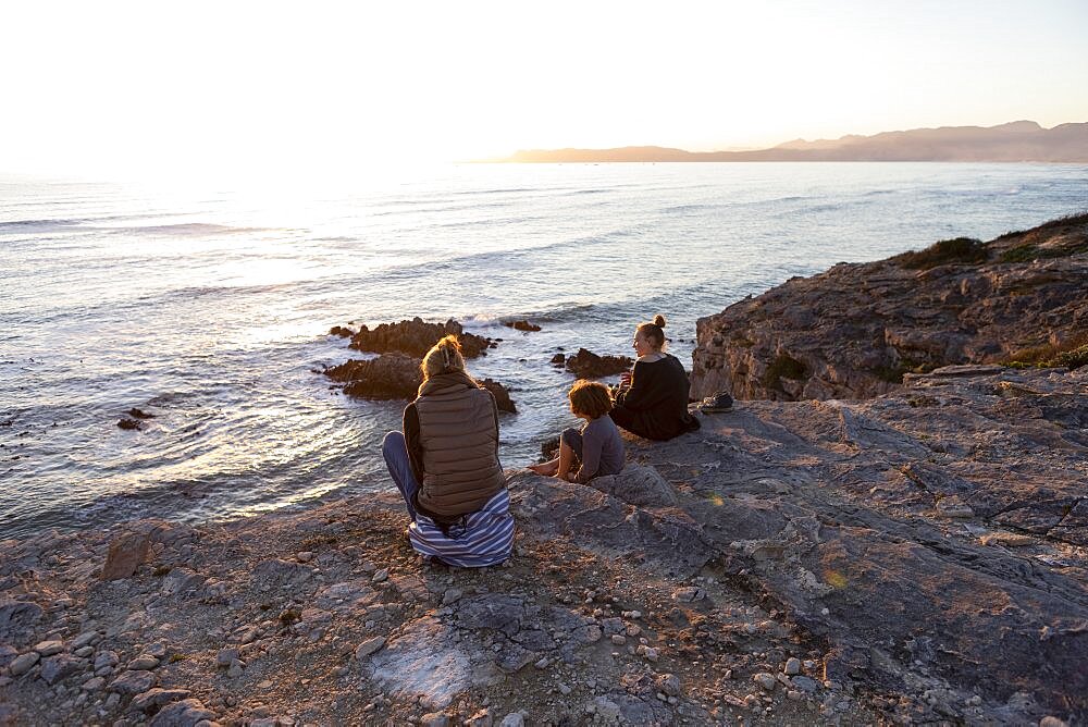 A family, mother and two children sitting watching the sunset over the ocean, Walker Bay Nature Reserve, South Africa