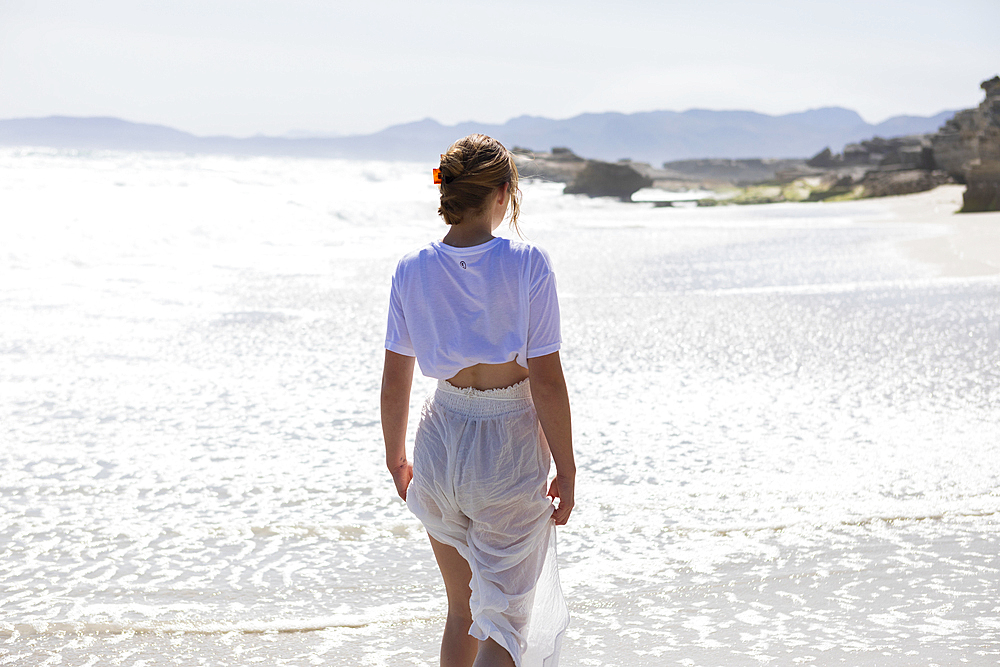 Teenage girl alone on a sandy beach in South Africa by the water's edge looking at the coastal view
