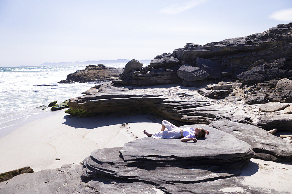 Teenage girl lying on her back on rocks above a sandy beach.