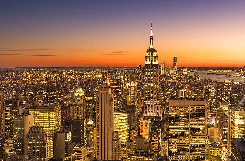 Empire state building rising above Manhattan skyline at sunrise or sunset.