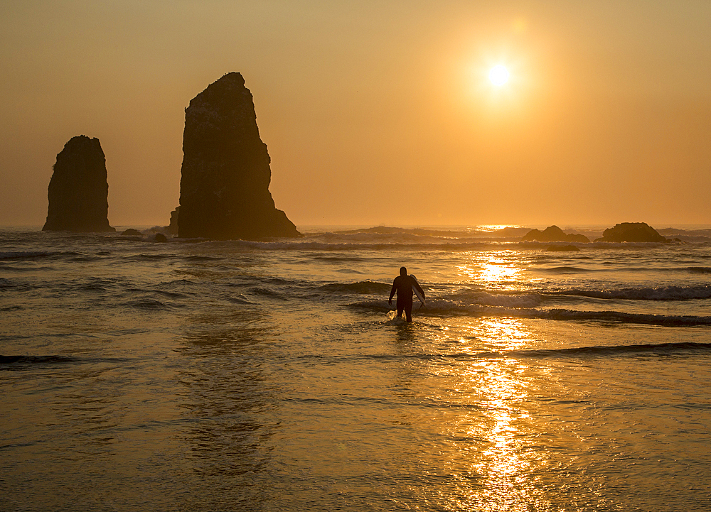 Surfer walking out of the sea with sun setting behind rock in waves on beach.