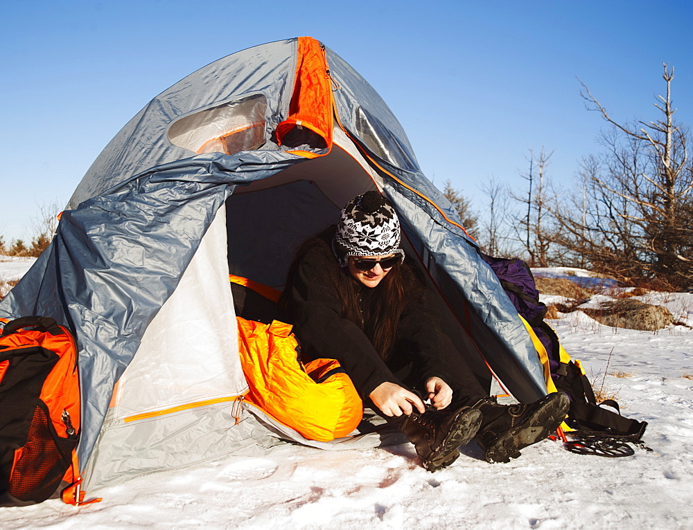 Woman sitting in tent in snowy landscape lacing up snow boots, Kingston, Ontario, Canada