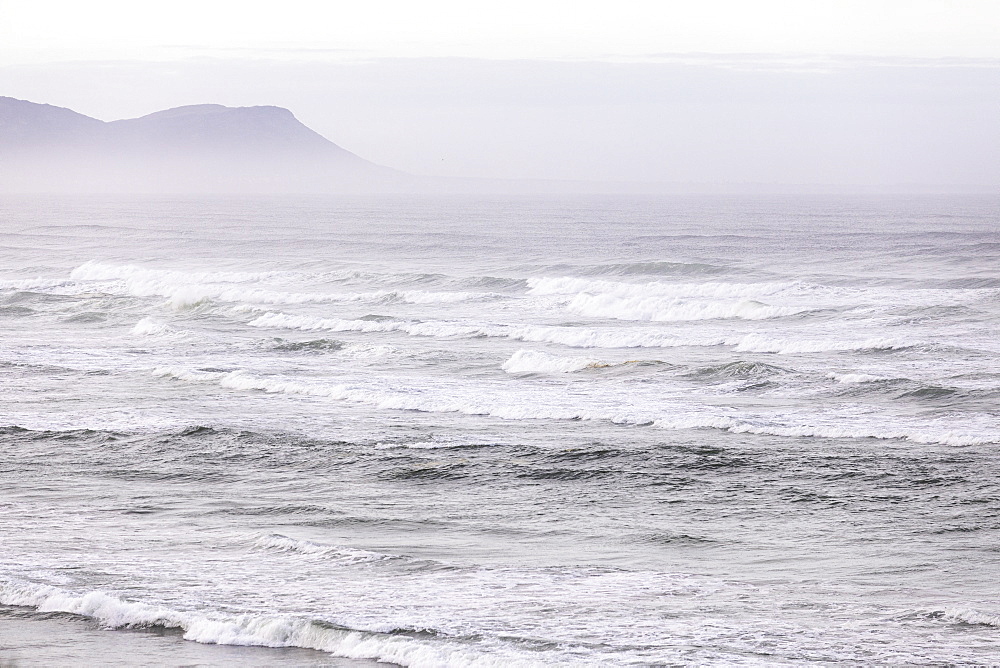 View over a sandy beach, mist rising and waves breaking on the Atlantic coastline, Voelklip Beach, Western Cape, South Africa