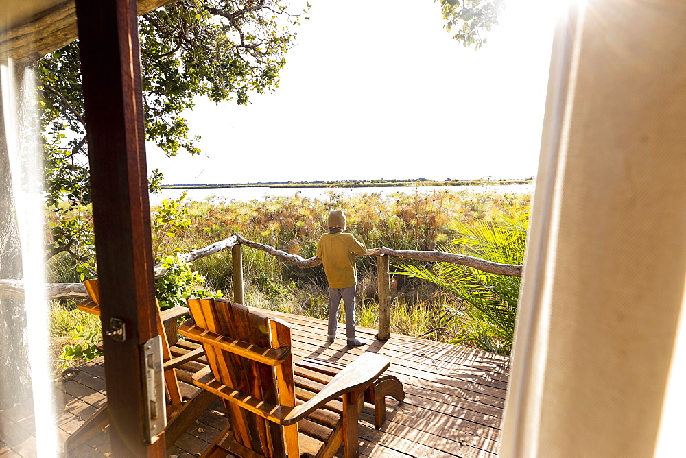 Young boy on wooden deck looking out over marshlands, Okavango Delta, Botswana