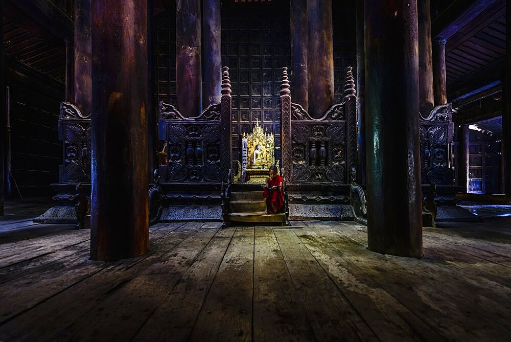 A Buddhist temple with wooden pillars and carved screens, a golden thrine and a young monk seated on the steps.