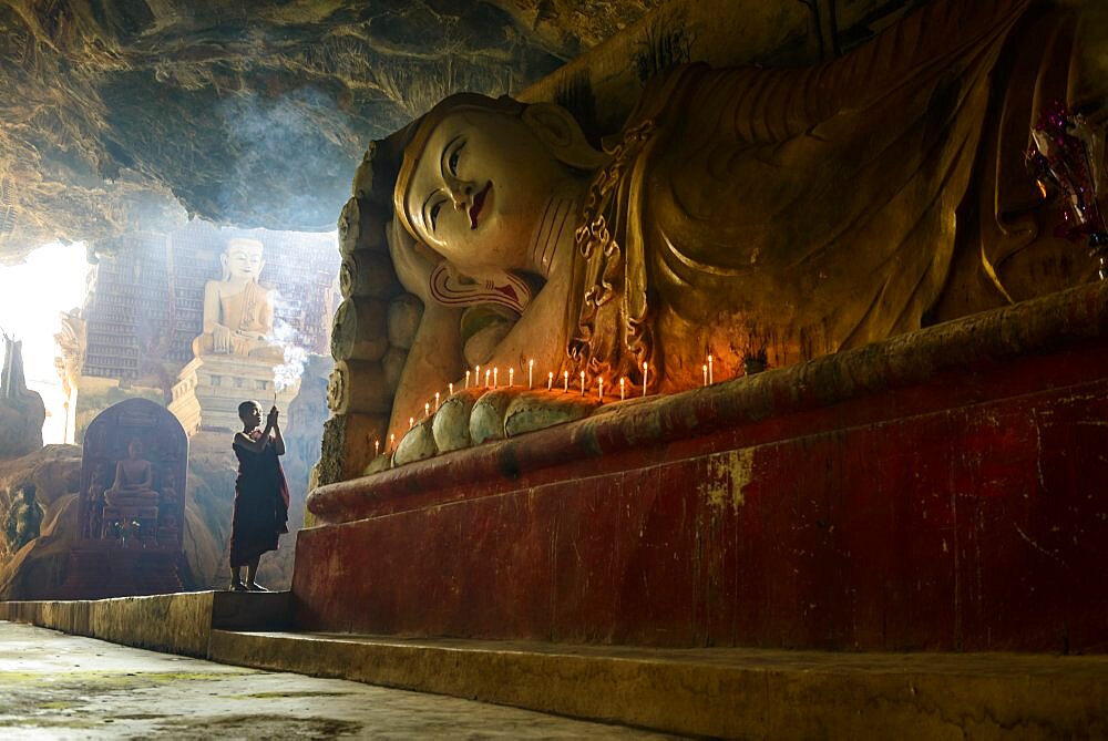 A cave temple, a large reclining Buddha statue, lit candles and a monk praying.