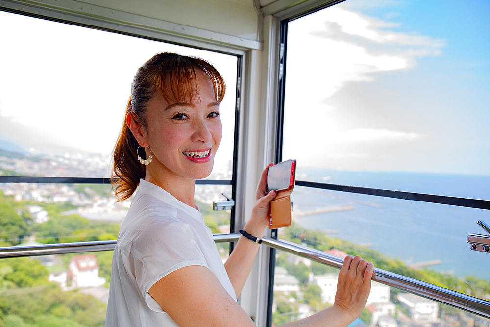 A mature Japanese woman using her mobile phone to take pictures from a cable car cabin of the city and landscape below.