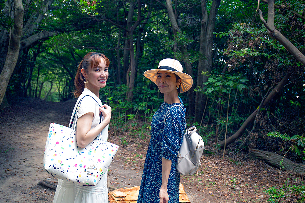 Two mature Japanese women in a park, carrying bags, on a day out, heads turned to smile at the camera.