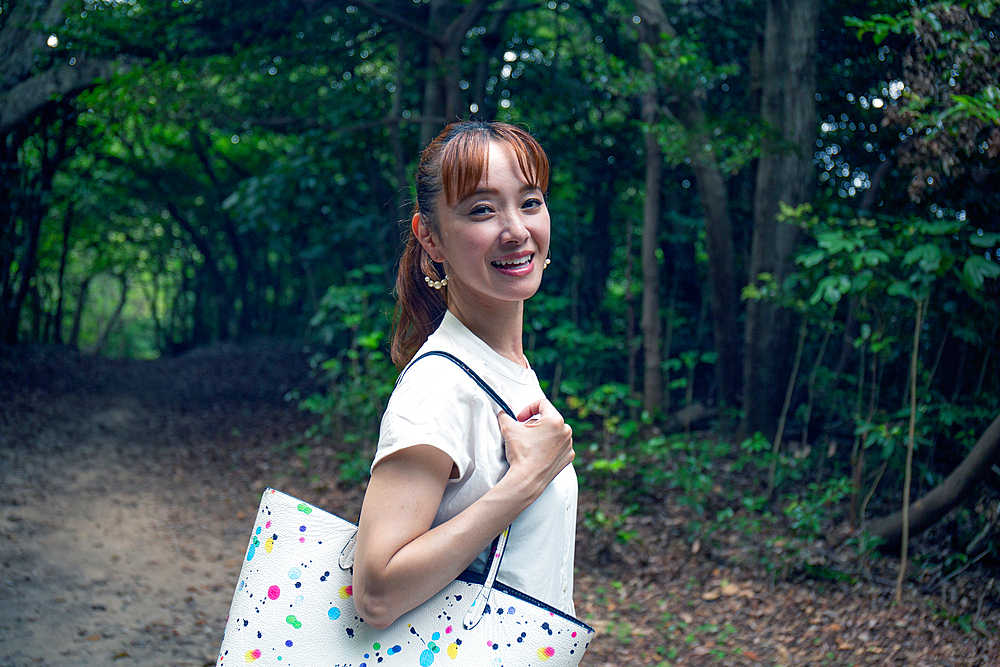 A mature Japanese woman outdoors in a park, head turned smiling at the camera.