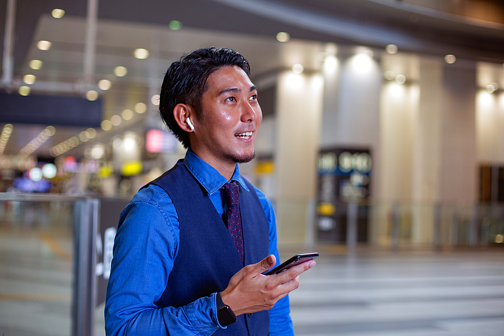 A young businessman in the city, standing looking around, holding his mobile phone.