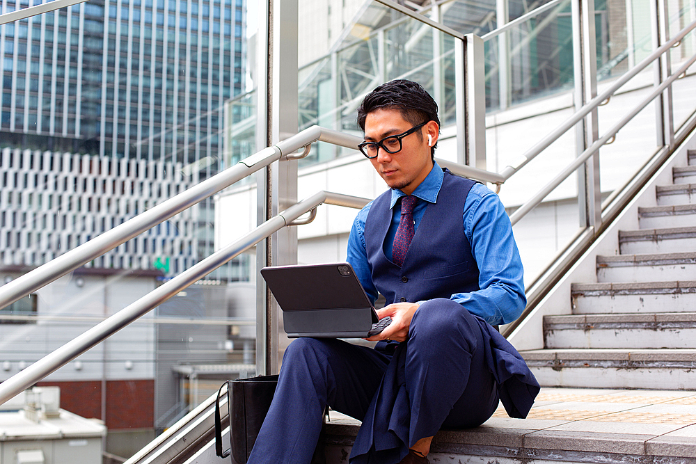 A young businessman in the city, on the move, seated on the stairs by a window, using a laptop.