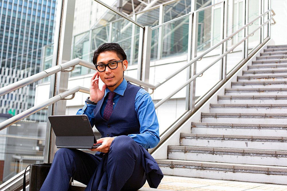 A young businessman in the city, on the move, sitting on stairs by a window on the phone, laptop open.